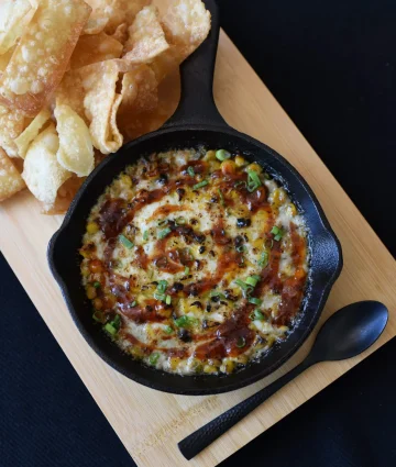 Golden baked cheese dip in cast iron skillet garnished with green onions, served with crispy potato chips on wooden board.