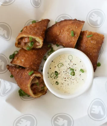 Golden-brown bacon-wrapped appetizers garnished with green onions, served with creamy dipping sauce for foodservice.