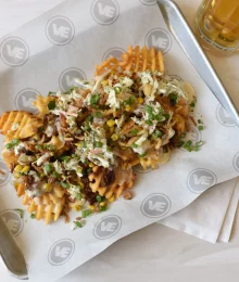 Golden waffle fries topped with melted cheese, crispy bacon, corn, and green onions on white paper for foodservice.