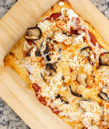 Golden-baked flatbread pizza topped with sautéed mushrooms, crispy bacon, melted cheese, and seasoning spices on wooden board.