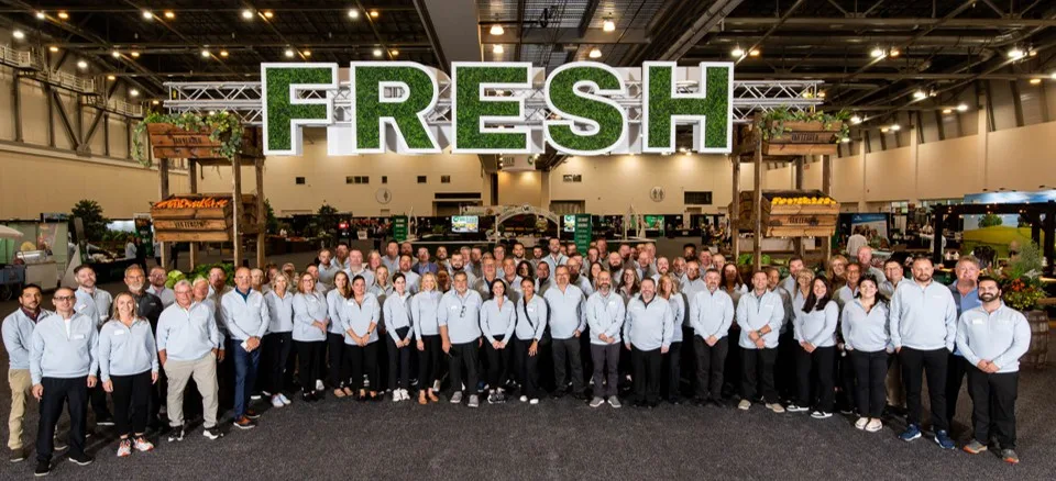 Large group of food industry professionals in white shirts standing together under Fresh signage at a food trade show or conference.